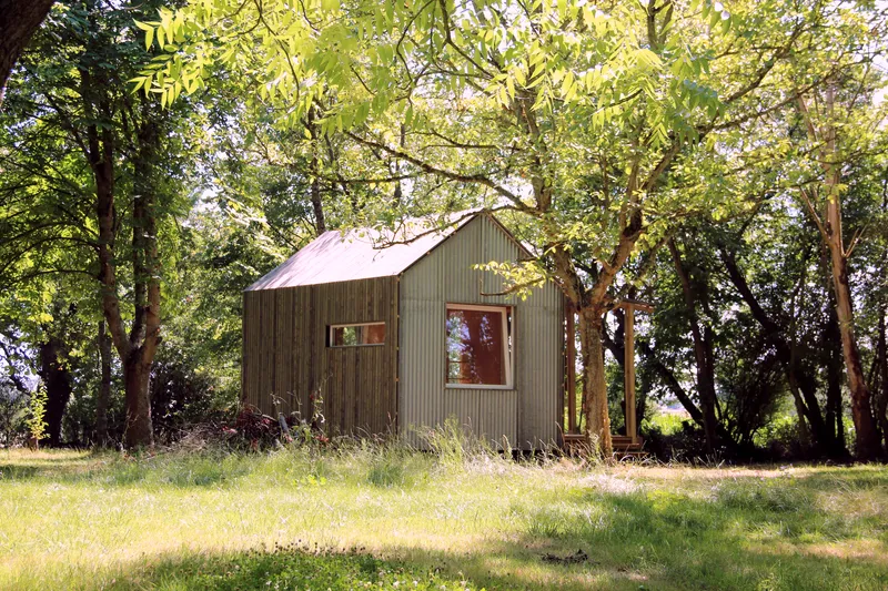 Gable-roofed cabin clad in corrugated metal and vertical timber siding nestled among mature trees in dappled sunlight