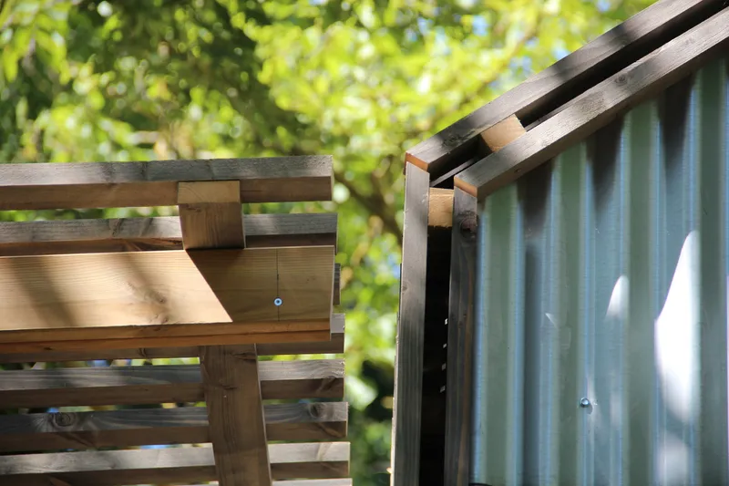 Detail of timber post and beam pergola structure meeting the corrugated metal wall in dappled sunlight