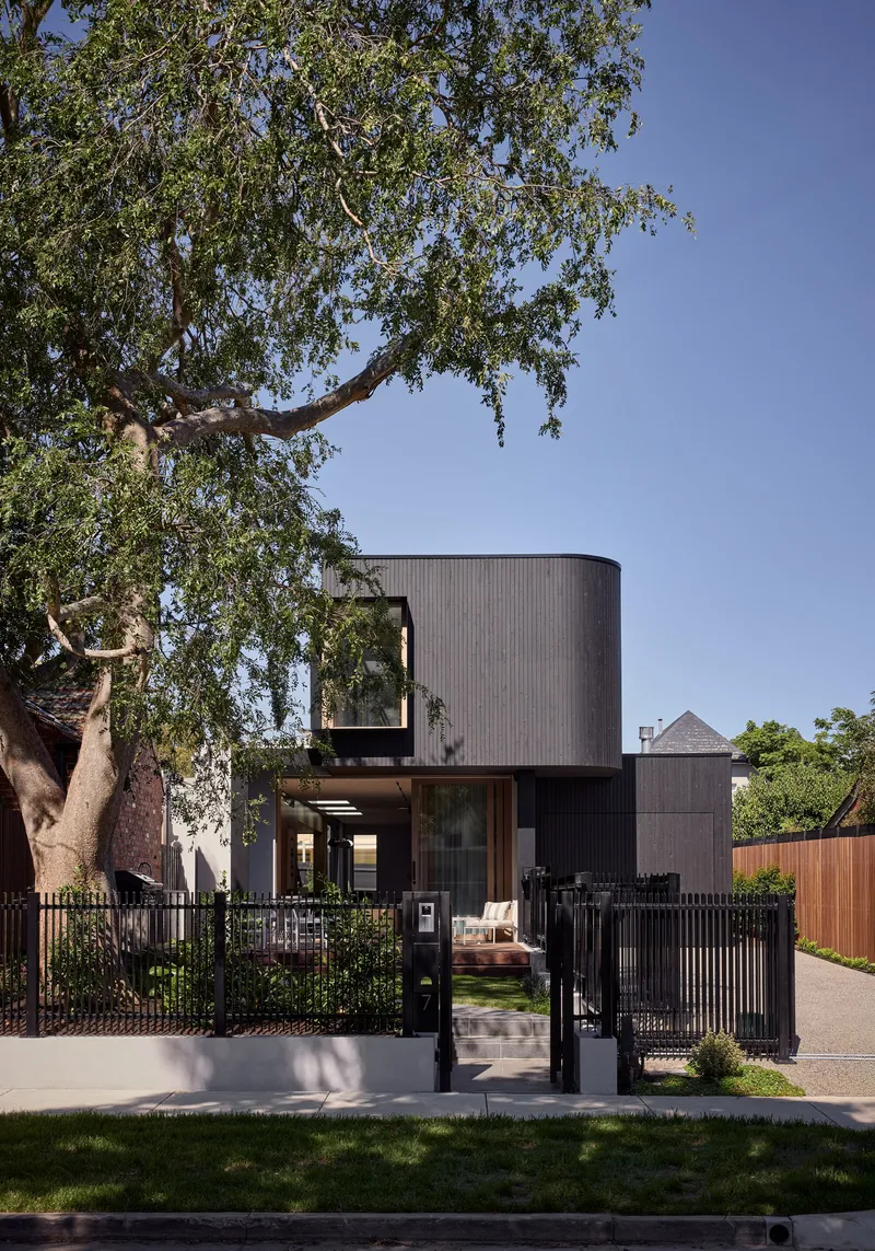 Street facade with black steel fence and mature tree shading the cylindrical volume and glazed lower level