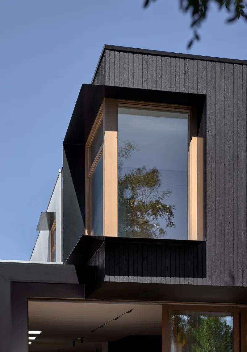 Cantilevered bay window with black metal frame and timber lining projecting from the grey ribbed facade at dusk