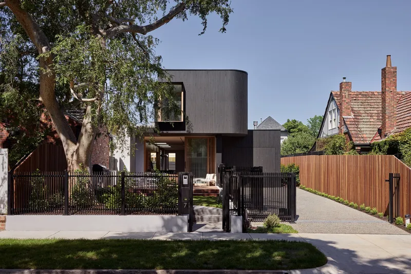 Street view of the black corrugated metal facade with vertical slat fencing and a mature tree overhead