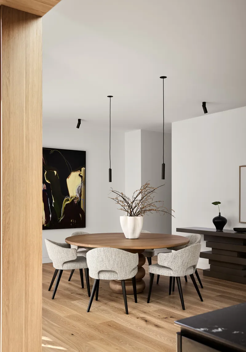Round dining table with upholstered chairs beneath suspended pendant lights in a white-walled room