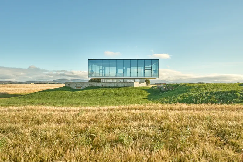Glass volume cantilevered above a grass-covered berm in a golden wheat field under afternoon light