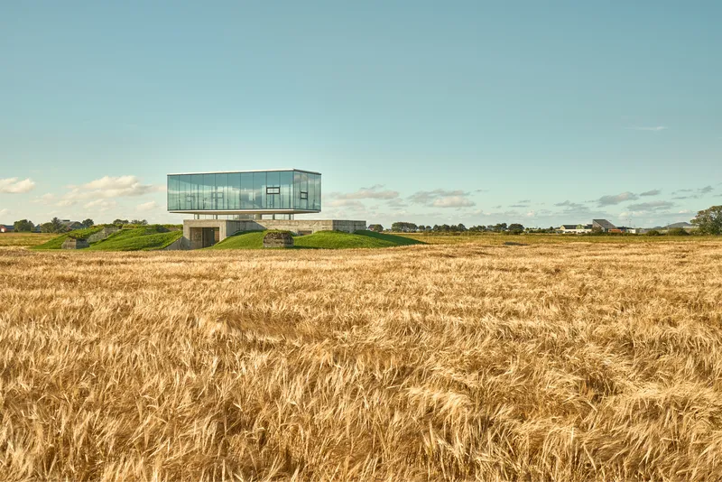 Elevated glass pavilion on concrete supports rising from a harvested wheat field