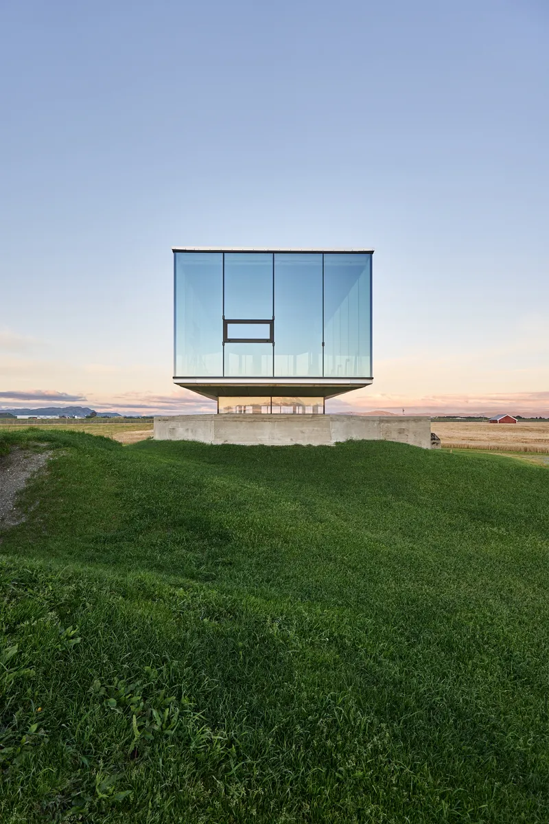 Front elevation of the glass box resting on concrete pilotis at dusk with manicured lawn below