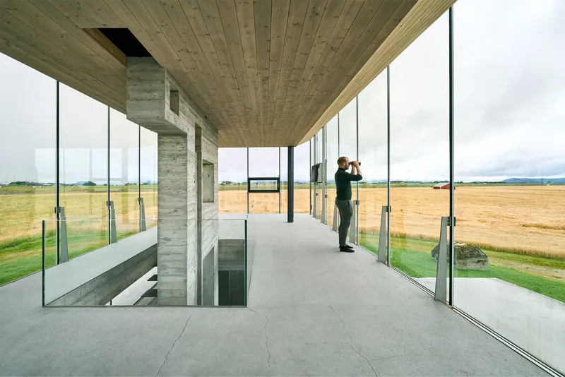 Interior corridor with timber plank ceiling and floor-to-ceiling glass overlooking farmland with a figure standing