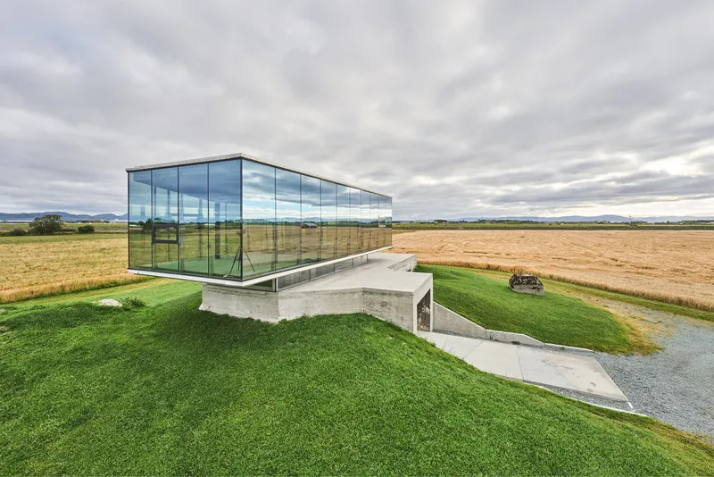 Glass pavilion cantilevered over a concrete base partially embedded in a grass-covered berm overlooking harvested fields