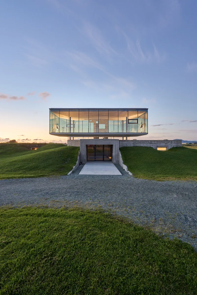 Frontal view of the glass volume cantilevered above a recessed concrete entrance at dusk