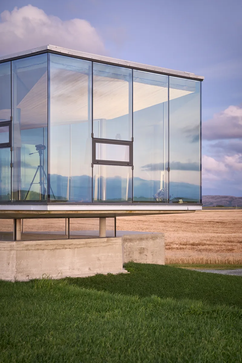 Corner detail of the glass pavilion with frameless glazing reflecting clouds above the open landscape