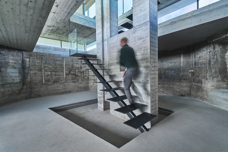 Board-formed concrete interior with a black steel staircase wrapping a column as a figure ascends