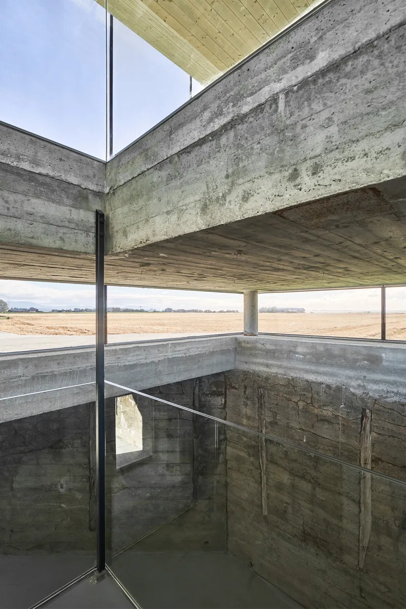 Corner glazing junction between board-formed concrete walls and timber ceiling with view to fields beyond