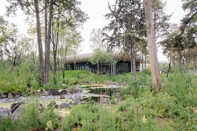 Glass-walled structure on a planted berm overlooking a natural pond with rocks and wildflowers