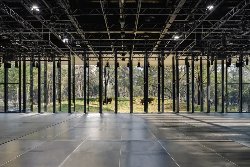 Interior view of exposed black structural grid ceiling and floor-to-ceiling glass overlooking trees