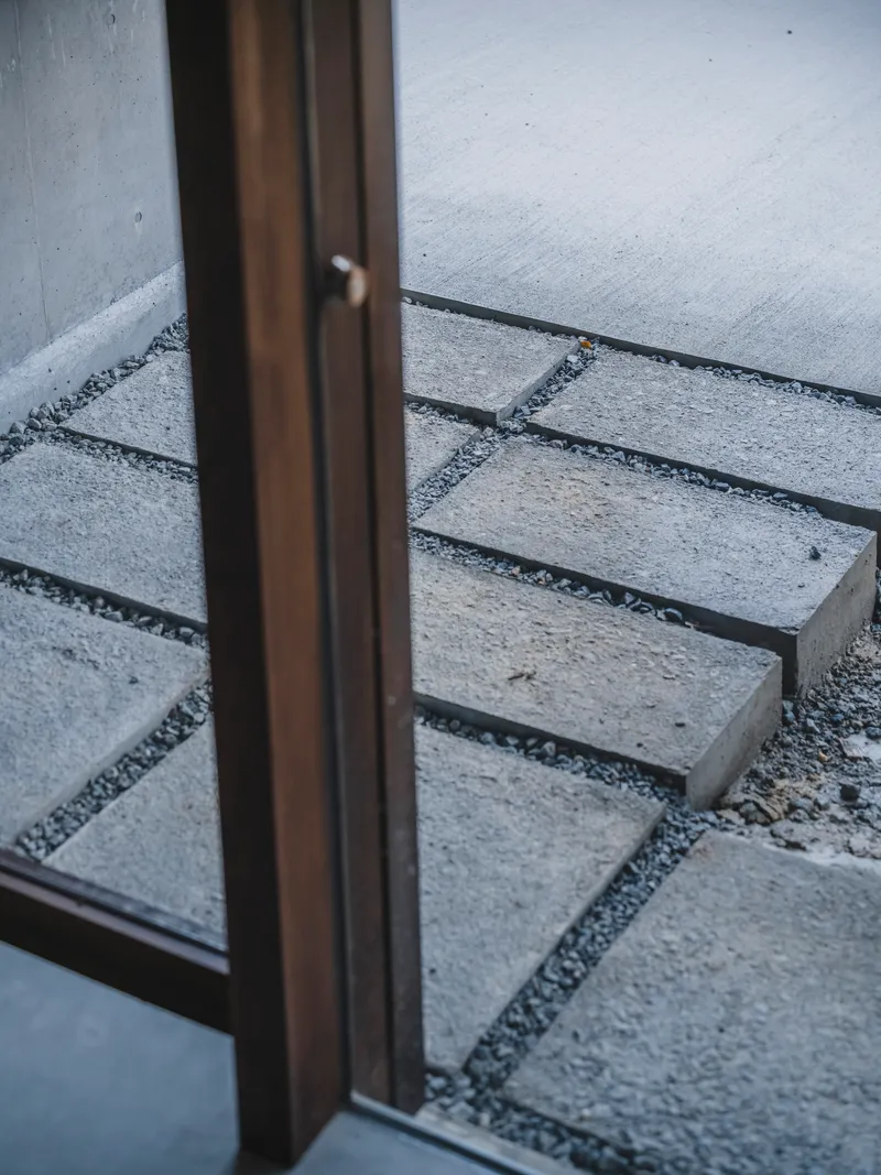Detail of bronze door frame meeting concrete paving slabs set in gravel at threshold