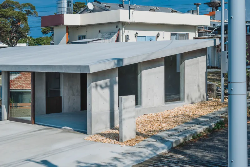 Concrete volume with flat overhang sheltering a recessed glazed opening above a gravel yard in daylight