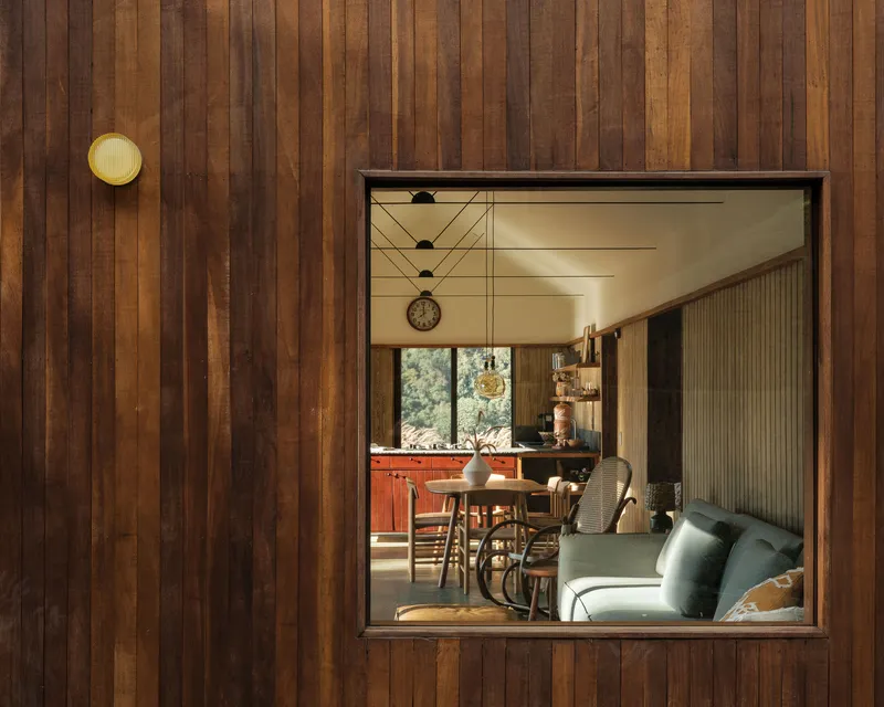Square window framed in vertical timber cladding reveals the dining area with pendant lights and a sofa