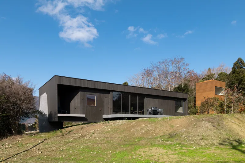 Dark timber-clad volume with cantilevered terrace perched on a grassy hillside slope under a blue sky