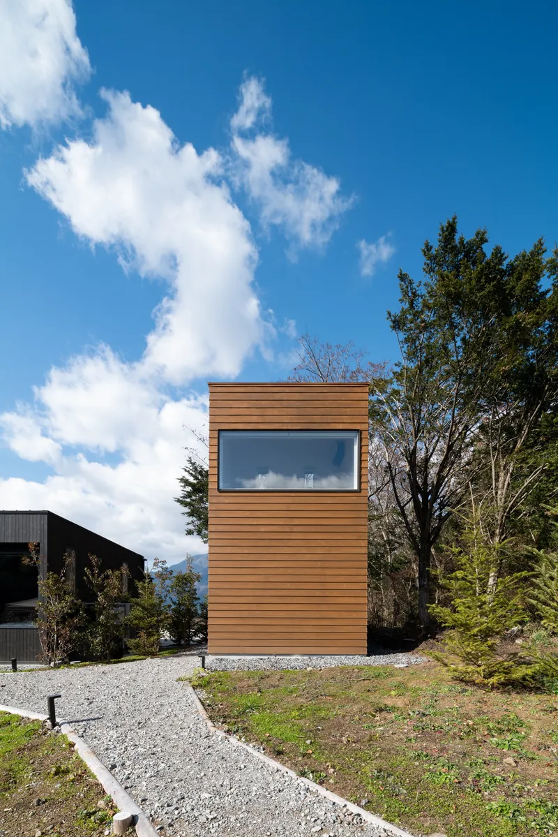 Freestanding cedar-clad tower with horizontal window band set on a gravel base beneath scattered clouds