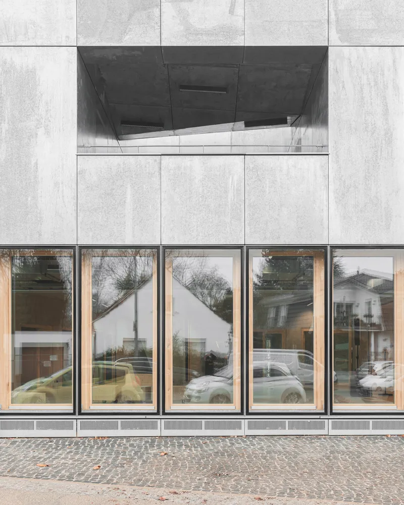 Facade detail showing concrete panel grid, horizontal glazing band with timber frames reflecting parked cars and trees