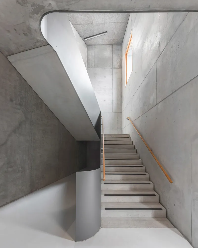Concrete stairwell with curving white balustrade and timber handrail under natural skylight