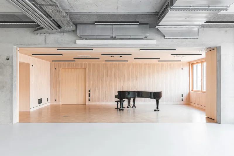 Practice room with grand piano enclosed by pale timber walls beneath exposed concrete ceiling and ductwork
