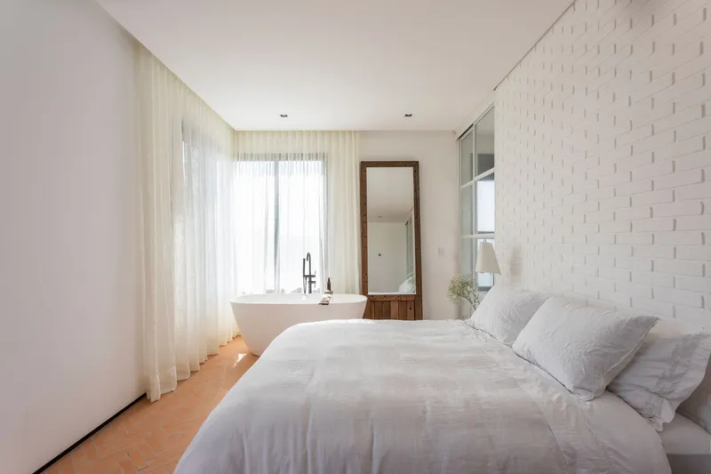 Bedroom with freestanding bathtub beside floor-to-ceiling windows draped in white sheer fabric