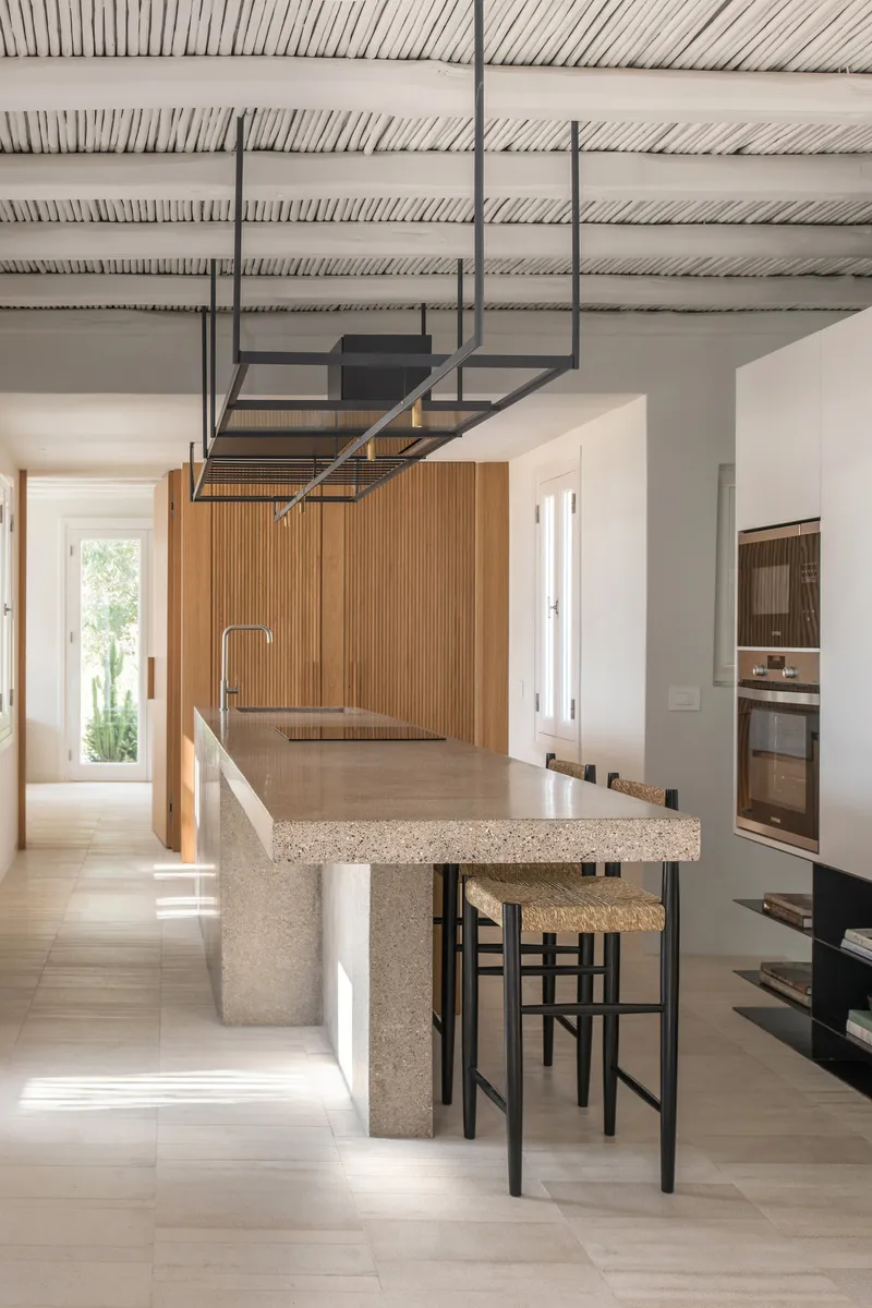 Kitchen island with terrazzo countertop and black metal stools beneath suspended steel shelving and timber ceiling