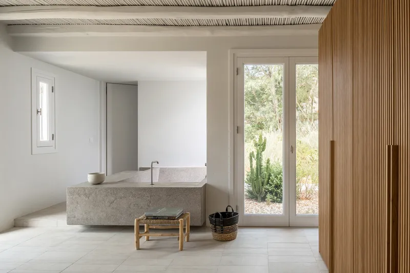 Bathroom with freestanding terrazzo tub, exposed timber ceiling beams, and glazed doors to planted courtyard