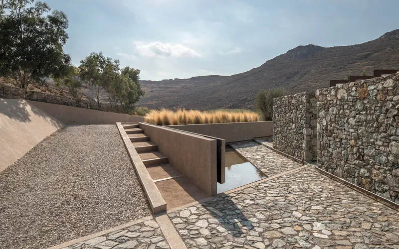Sunken courtyard pool framed by gravel paths and stone walls beneath hillside and ornamental grasses
