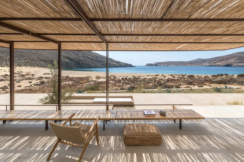 Long dining table under woven reed ceiling with view across coastal terrain to bay