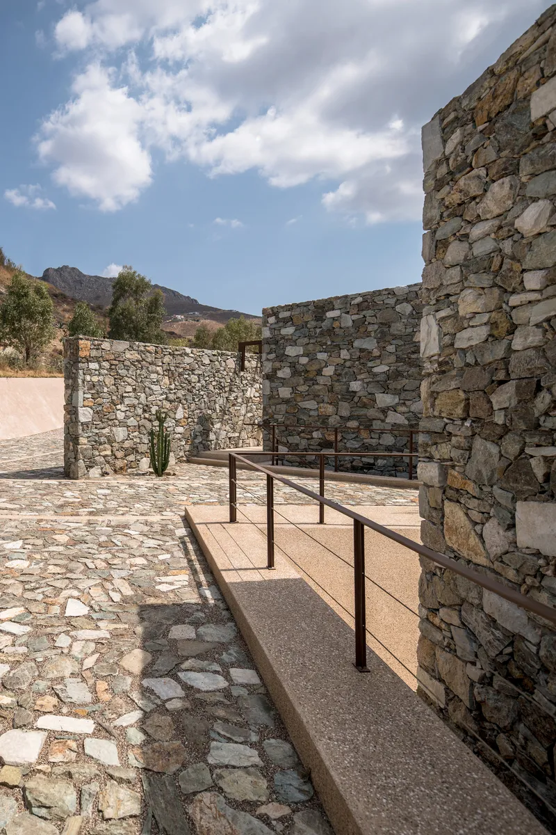 Stone-paved courtyard with metal-railed ramp between textured masonry walls under afternoon clouds