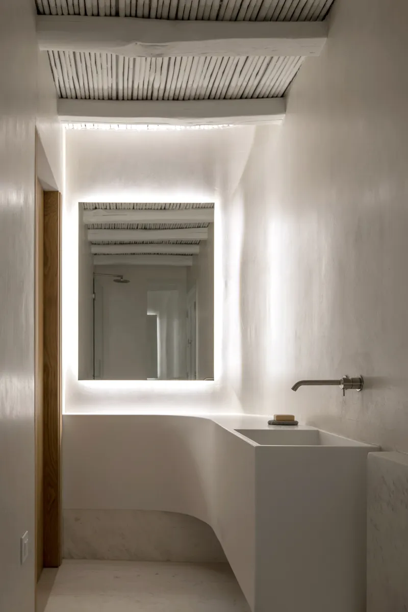Bathroom with backlit mirror and white plaster vanity under exposed timber slat ceiling and indirect cove lighting