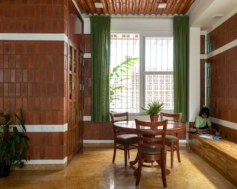 Dining area with brick tile walls and green curtains framing a window where a person sits reading