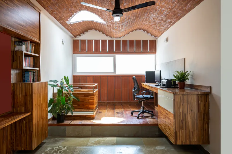 Home office with vaulted brick ceiling, timber desk, and potted plants beneath a horizontal window