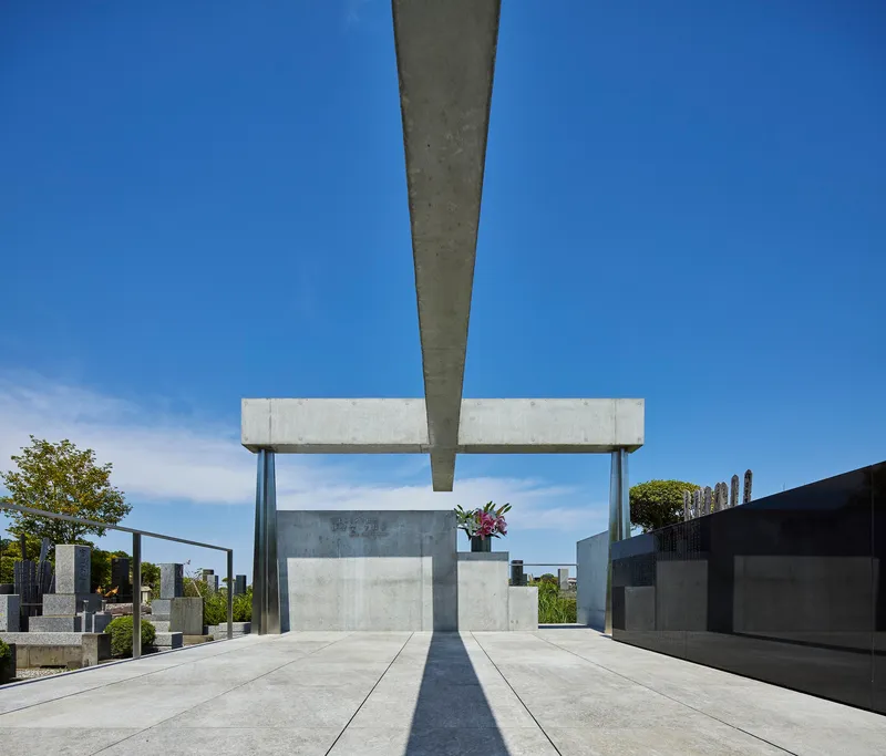 View along concrete plaza with central monument beneath cantilevered beam casting sharp shadows at midday