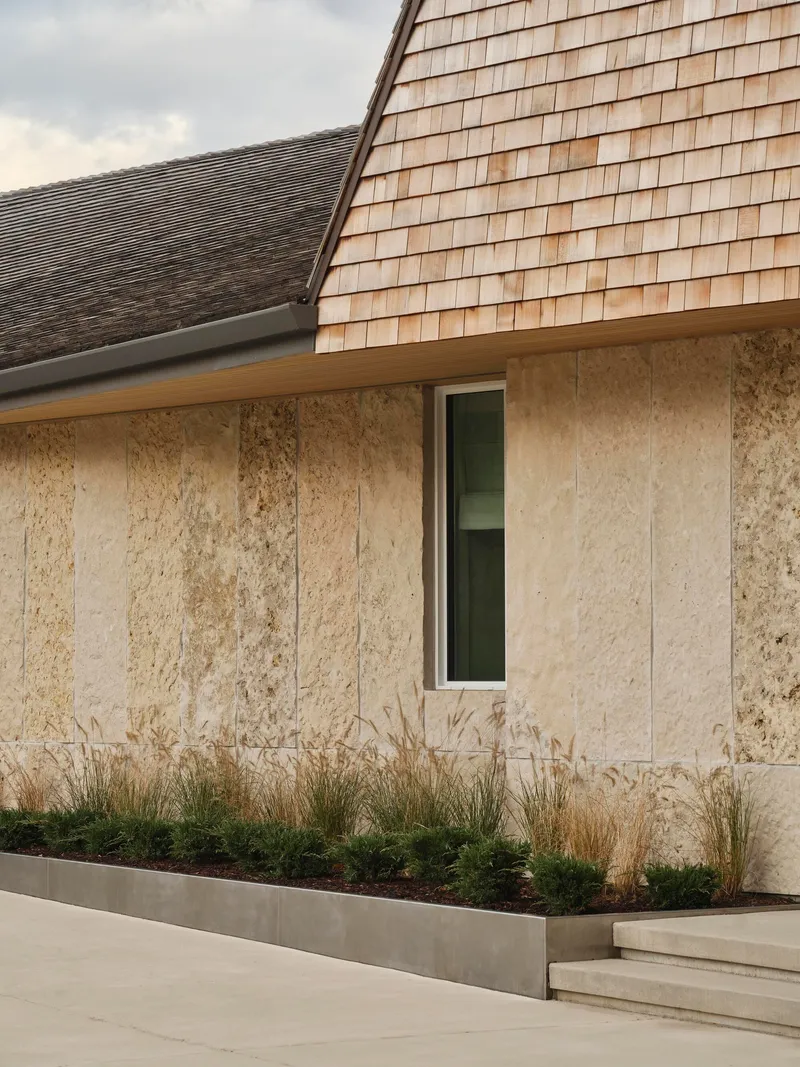 Exterior facade with textured stone panels and cedar shingle roof above a planted bed with dried grasses