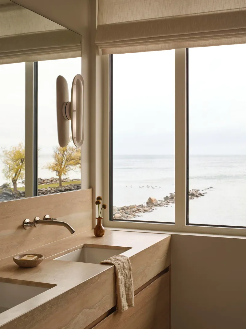 Bathroom vanity in pale stone with wall-mounted faucet facing windows overlooking the waterfront
