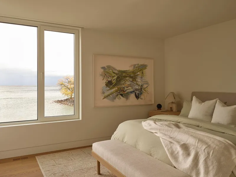 Bedroom with corner window framing a lone tree and distant water under overcast sky