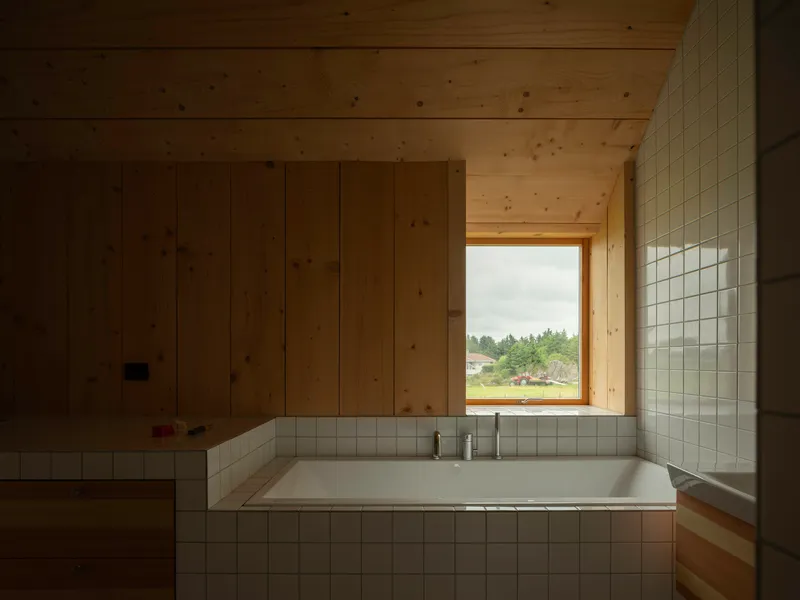 Recessed white-tiled bathtub beneath a timber-framed window overlooking distant forest and shoreline