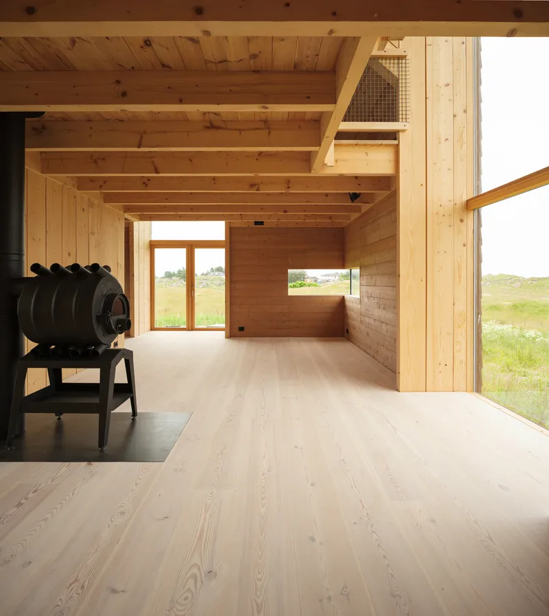Interior view of timber room with exposed beams and a black wood stove overlooking grassy landscape