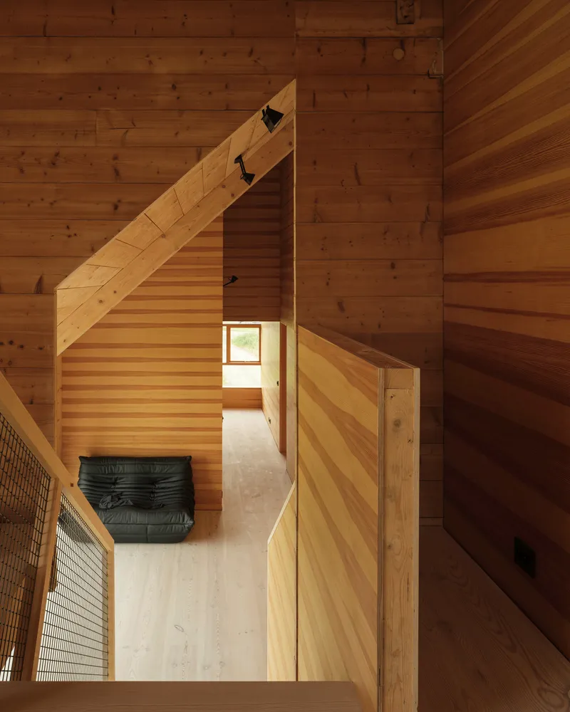 View down a double-height timber-lined corridor with diagonal wood slat stair rising alongside a black leather bench