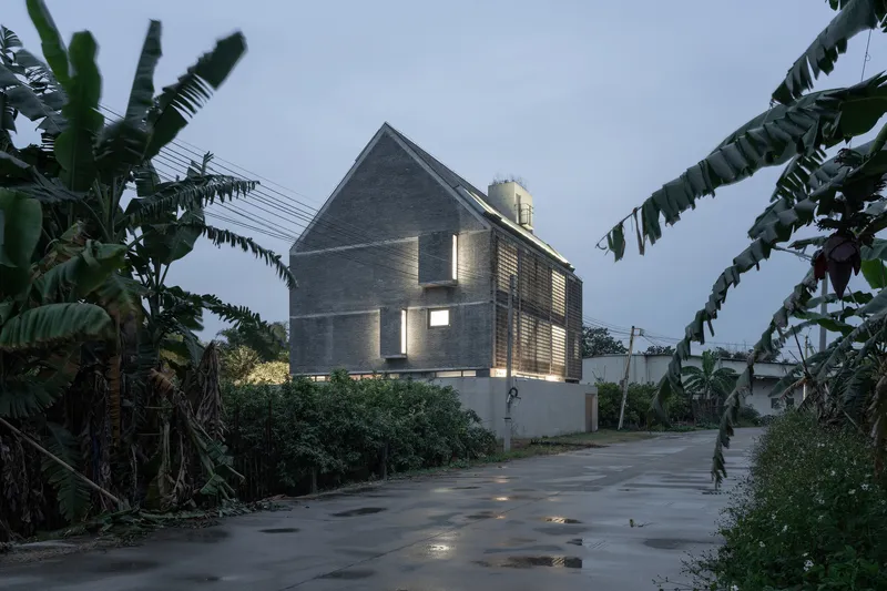 Gable-roofed concrete house with illuminated slit windows viewed from street flanked by banana plants