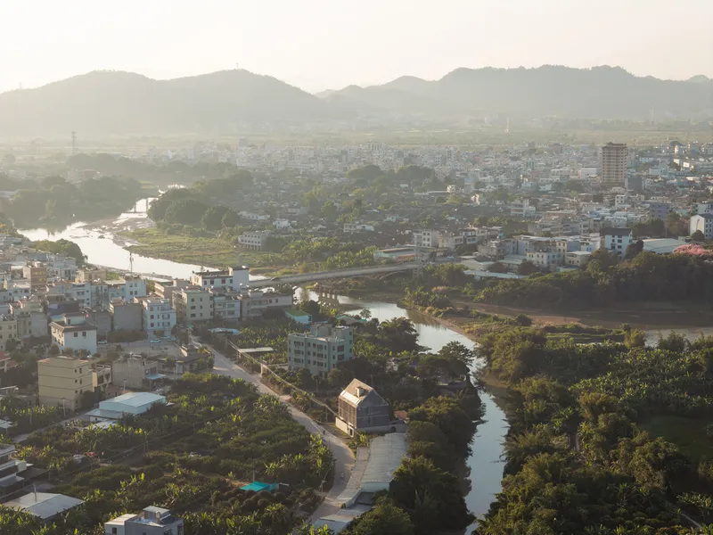 Aerial view of a gable-roofed structure nestled along a winding river with mountains in the distance at dusk
