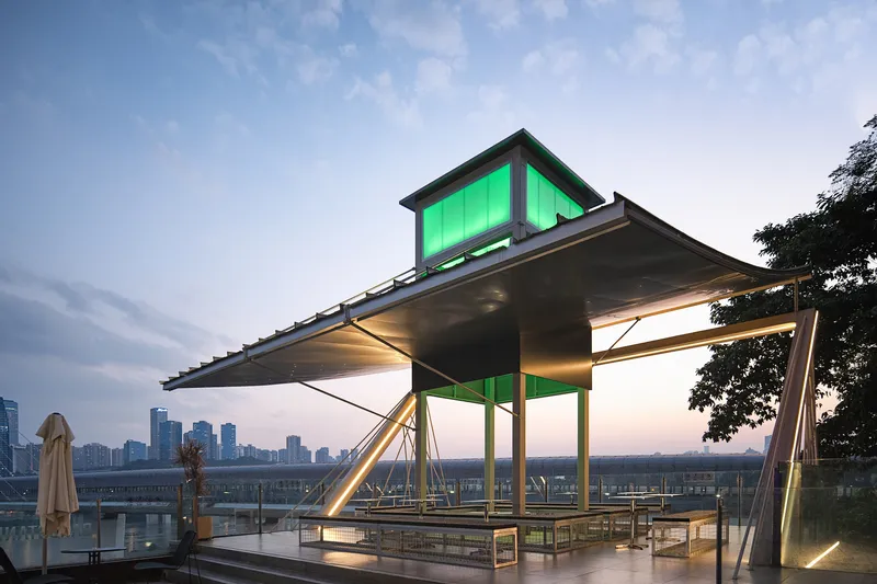 Rooftop pavilion with green glowing upper volume and cantilevered corrugated metal roof at dusk
