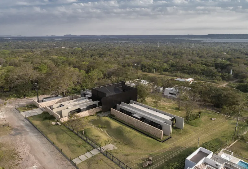 Aerial view of the black metal volumes and stone walls arranged across a grassy clearing