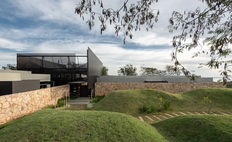 Entrance path between stone retaining walls and grassed berms beneath corrugated metal volumes and eucalyptus branches