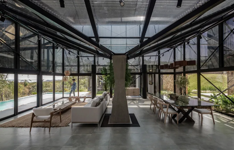 Glass-walled living area with exposed steel structure, central concrete fireplace and corrugated metal ceiling