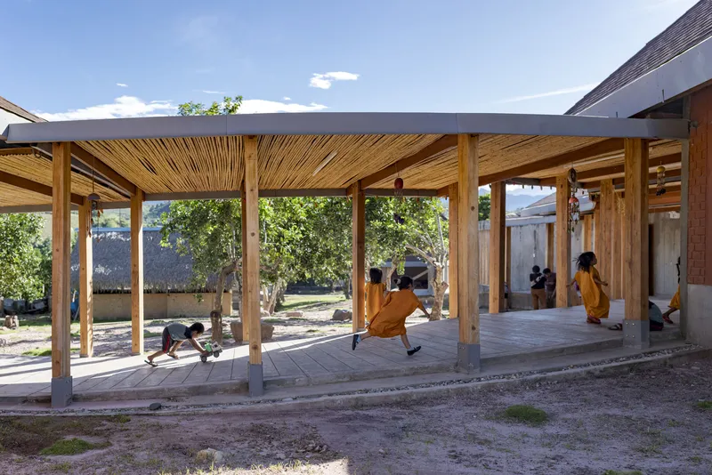 Open timber colonnade with slatted wood ceiling and concrete floor where children play beneath the shade