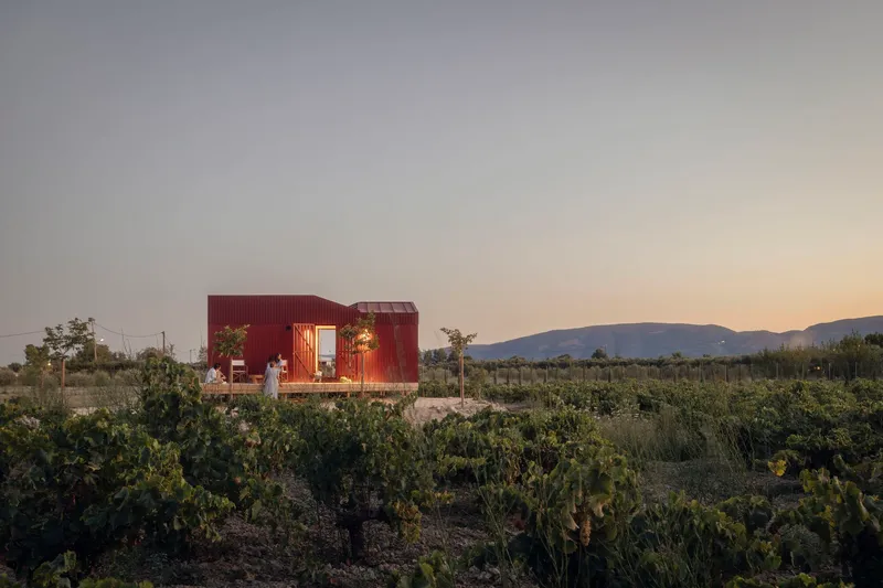 Red corrugated metal pavilion with open side opening set in a scrubland field at dusk