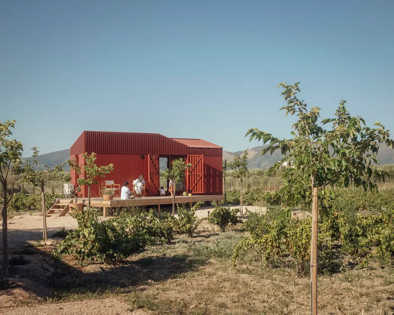 Timber deck with seated figures in front of the red metal structure and young orchard